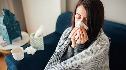 Erkältete Frau auf dem Sofa mit Decke um die Schultern putzt sich die Nase - Foto: iStock/evrim ertik