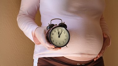 Frau hält sich den Babybauch und hat eine Uhr in der Hand - Foto: iStock/ jodiecoston
