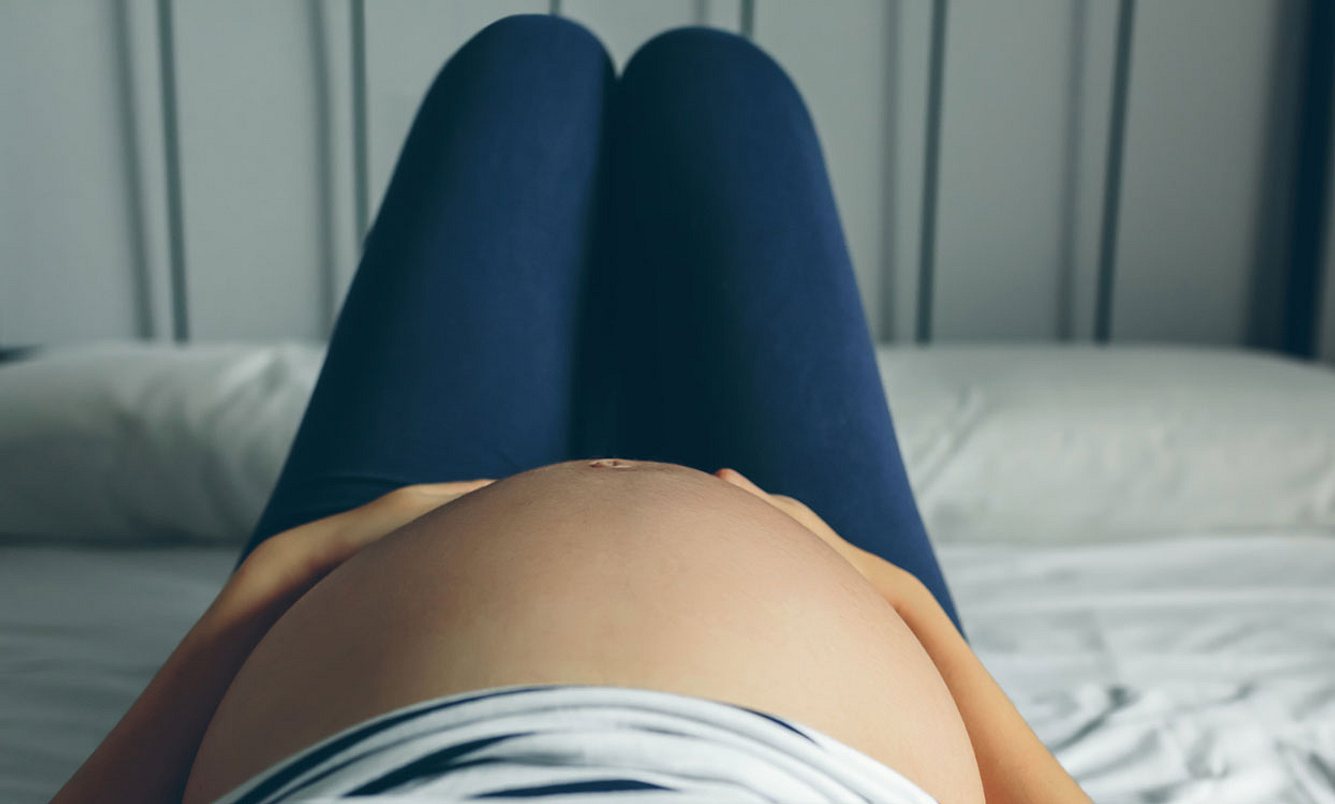 Schwangere liegt im Bett und hält Hände auf den Unterleib