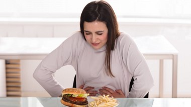 Frau hält sich den Bauch und sitzt vor Burger und Pommes - Foto: iStock/AndreyPopov