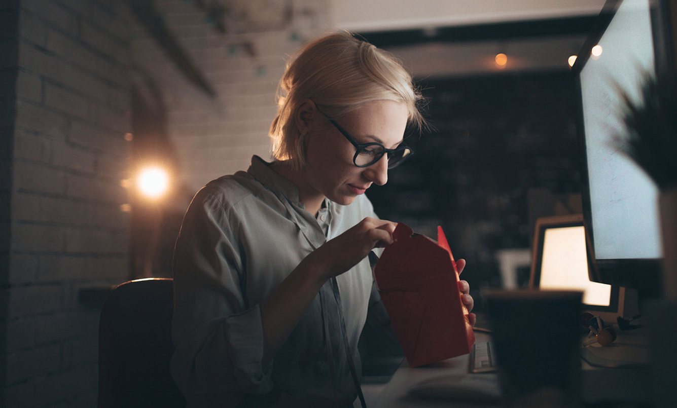 Eine Frau arbeitet am Computer und isst dabei ein Fertiggericht aus einer Pappbox