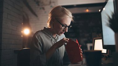 Eine Frau arbeitet am Computer und isst dabei ein Fertiggericht aus einer Pappbox - Foto: iStock/AleksandarNakic