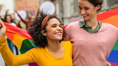 Zwei lachende Frauen laufen Arm in Arm mit und halten eine Regenbogenflagge - Foto: iStock/Ridofranz