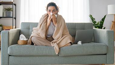Eine erkältete Frau sitzt auf dem Sofa und putzt ihre Nase - Foto: iStock_Choreograph