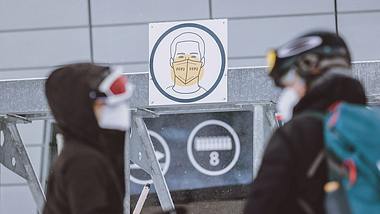 Zwei Skifahrer in Schwarz vor Hinweisschild auf Maskenpflicht - Foto: IMAGO / Eibner Europa