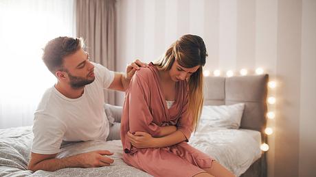 Eine Frau sitzt im Bett und hat Bauchschmerzen, ihr Mann tröstet sie - Foto: iStock/Jelena Danilovic