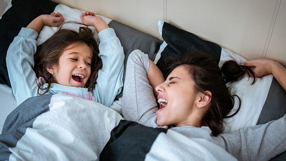 EIne Mutter liegt mit ihrer kleinen Tochter im Bett und beide gähnen - Foto: iStock/Johnce