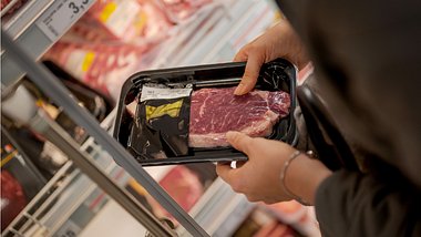 Jemand hält ein in Plastik eingeschweißtes Schweinesteak in der Hand vor dem Supermarkregal - Foto: istock_webphotographeer