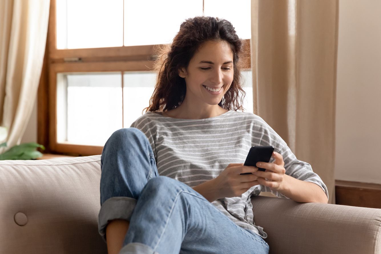 Lächelnde Frau sitzt auf dem Sofa und schaut auf ihr Smartphone