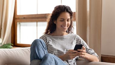 Lächelnde Frau sitzt auf dem Sofa und schaut auf ihr Smartphone - Foto: iStock-ID: 1207723294 fizkes
