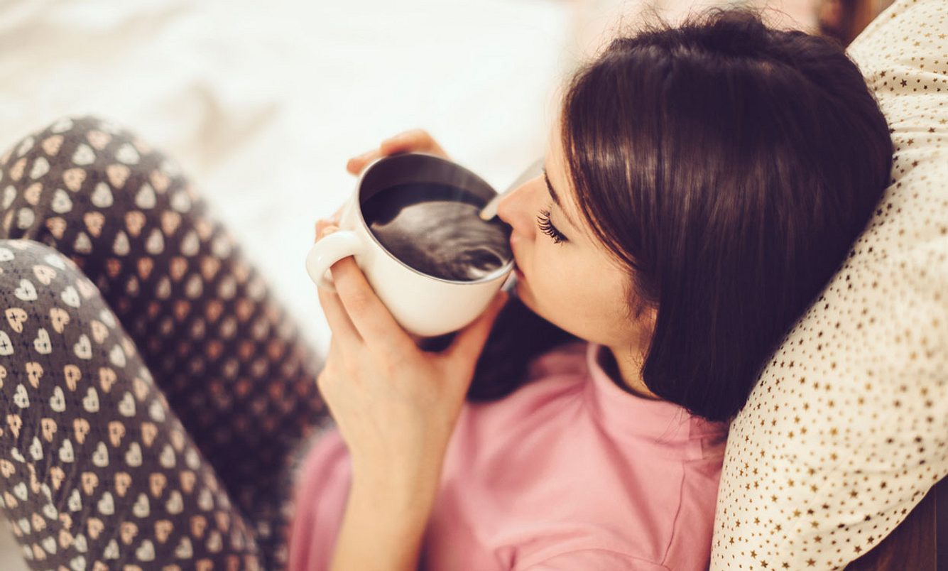tee-gegen-bauchschmerzen Frau sitzt im Bett und trinkt Tee gegen Bauchschmerzen