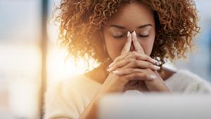 Eine gestresste Frau sitzt mit geschlossenen Augen am Computer - Foto: iStock/PeopleImages