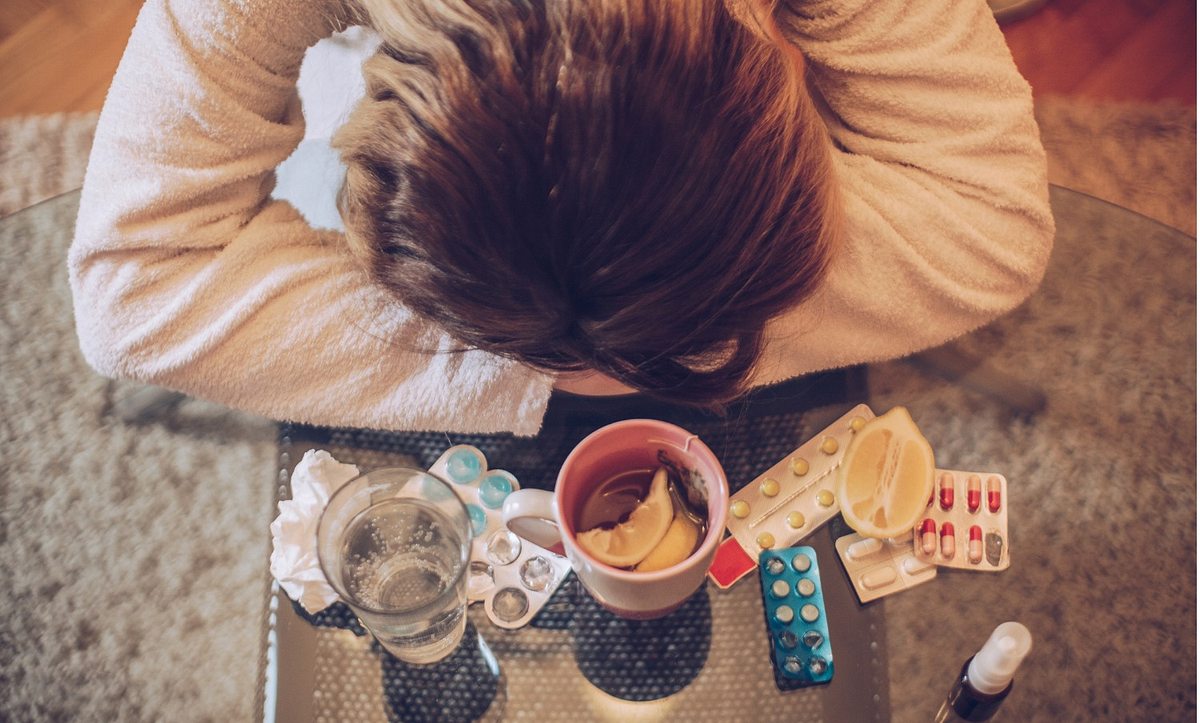 Frau liegt mit Kopf auf verschränkten Armen auf dem Tisch mit Medikamenten um sie herum