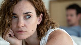 Frau sitzt von einem Mann abgewandt auf einem Bett und schaut nachdenklich. - Foto: iStock_Mindful Media