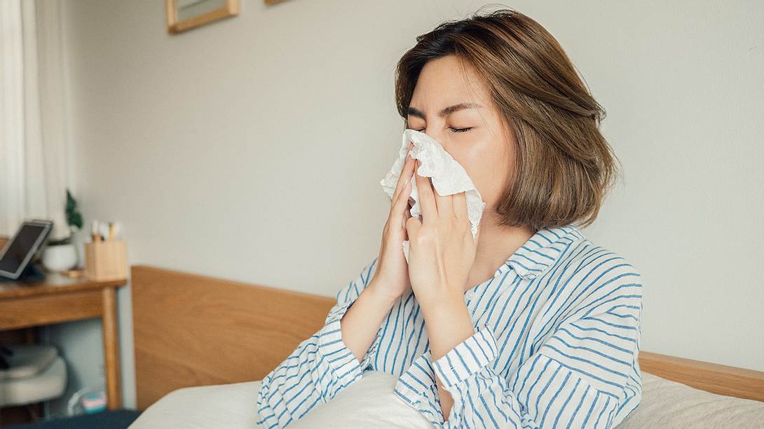 Eine Frau sitzt im Bett und putzt sich die Nase - Foto: iStock/staticnak1983