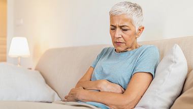 Eine ältere Frau sitzt auf dem Sofa und hält sich den Bauch - Foto: iStock/stefanamer