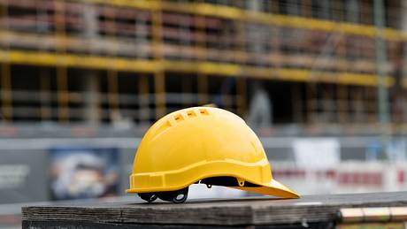 Gelber Bauarbeiter-Helm, im Hintergrund eine Baustelle mit Gerüsten - Foto: iStock/Cineberg Gelber Bauarbeiter-Helm, im Hintergrund eine Baustelle mit Gerüsten - Foto: iStock/Cineberg