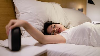 Junge Frau liegt im Bett und guckt müde auf den Wecker - Foto: iStock/rudi_suardi