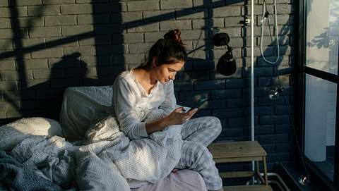 Eine Frau sitzt im Schlafanzug auf ihrem Bett und schaut auf ihr Smartphone - Foto: iStock/AleksandarNakic