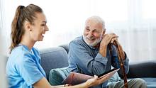 Pflegekraft und älterer Mann sitzen lächelnd auf einer Couch und schauen sich ein Bildband an. - Foto: iStock / Paperkites