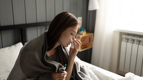 Frau sitzt auf dem Bett und putzt sich die Nase. - Foto: iStock/Niyaz_Tavkaev