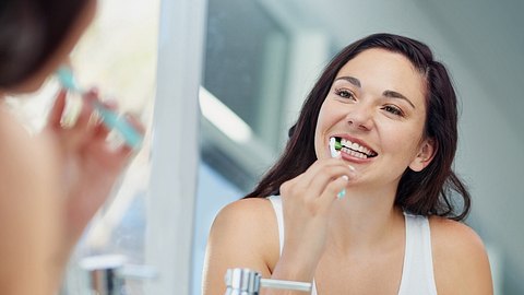 Eine Frau putzt sich die Zähne und schaut dabei in den Spiegel - Foto: istock_LaylaBird